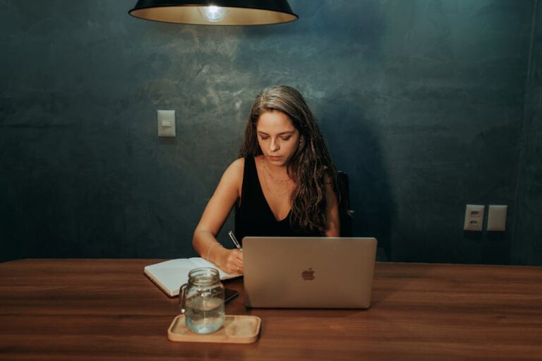 Focused woman writing notes while working on a laptop in a dimly lit room.
