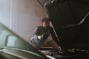A male mechanic checks under the hood of a car in a dimly lit garage, focusing on vehicle repair.