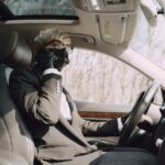 From below side view of unrecognizable woman in formal clothes and protective mask and gloves talking on cellphone while driving contemporary car