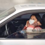 Female driver wearing warm outfit adjusting protective facial mask while sitting in auto with opened window during coronavirus pandemic in city and looking at camera