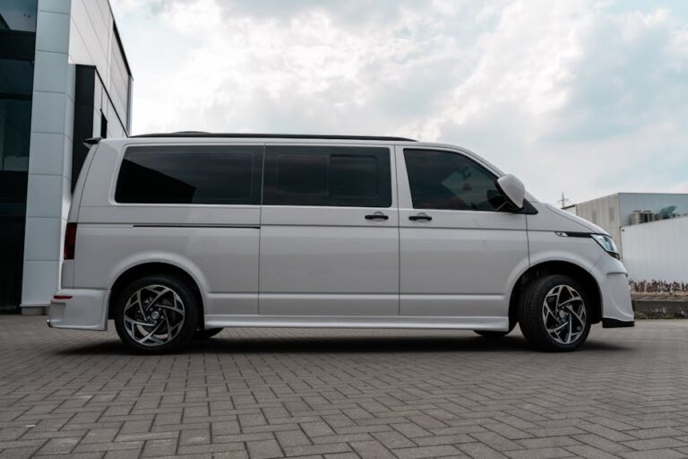 A modern white van parked outdoors on stone pavement with visible rims and side mirror.