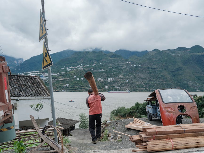 A man carries a wooden log by a riverside with mountains in the background.