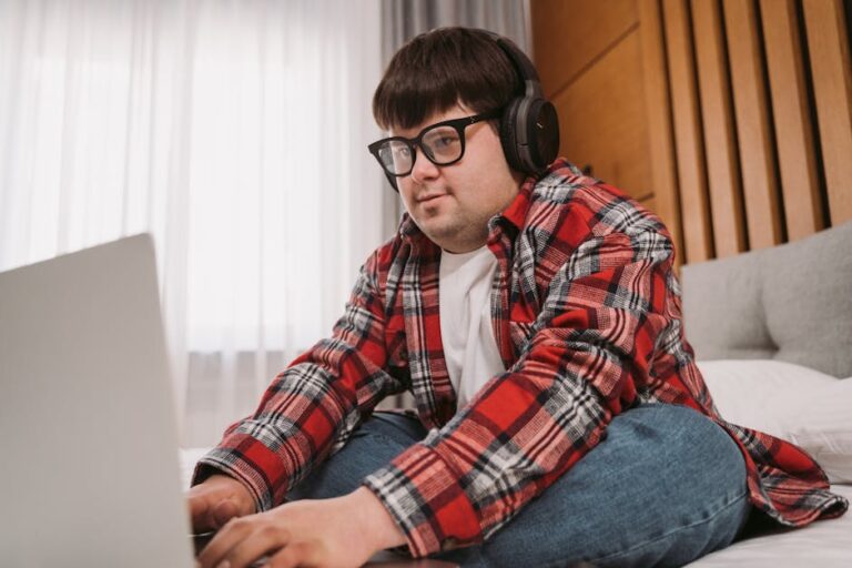 Young man wearing headphones and glasses using laptop on bed indoors, relaxed and focused.