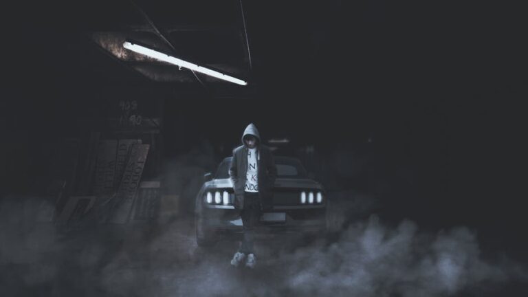 A hooded person stands confidently against a car in a foggy, dimly lit garage.