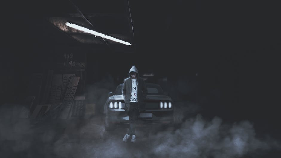 A hooded person stands confidently against a car in a foggy, dimly lit garage.