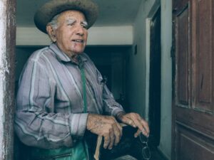 A senior man wearing a cowboy hat stands in a doorway, holding a book and glasses.