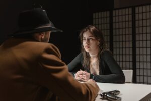 A woman faces a man in a hat during a tense interrogation scene at a table.
