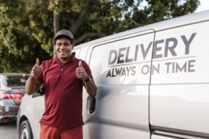 Happy deliveryman in maroon shirt giving thumbs up by delivery van outdoors.
