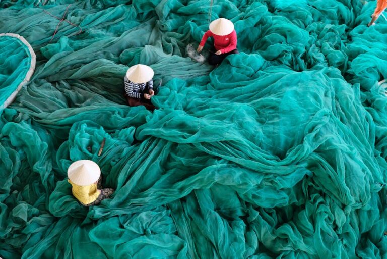 Aerial view of three people weaving turquoise fishing nets, showcasing traditional craftsmanship and textiles.