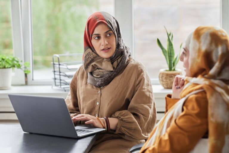 Two women in hijabs work together in a modern office, discussing at a laptop.