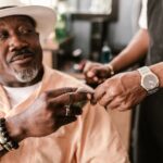 Senior man with a hat giving a tip to a young barber indoors, wearing bracelets and a watch.