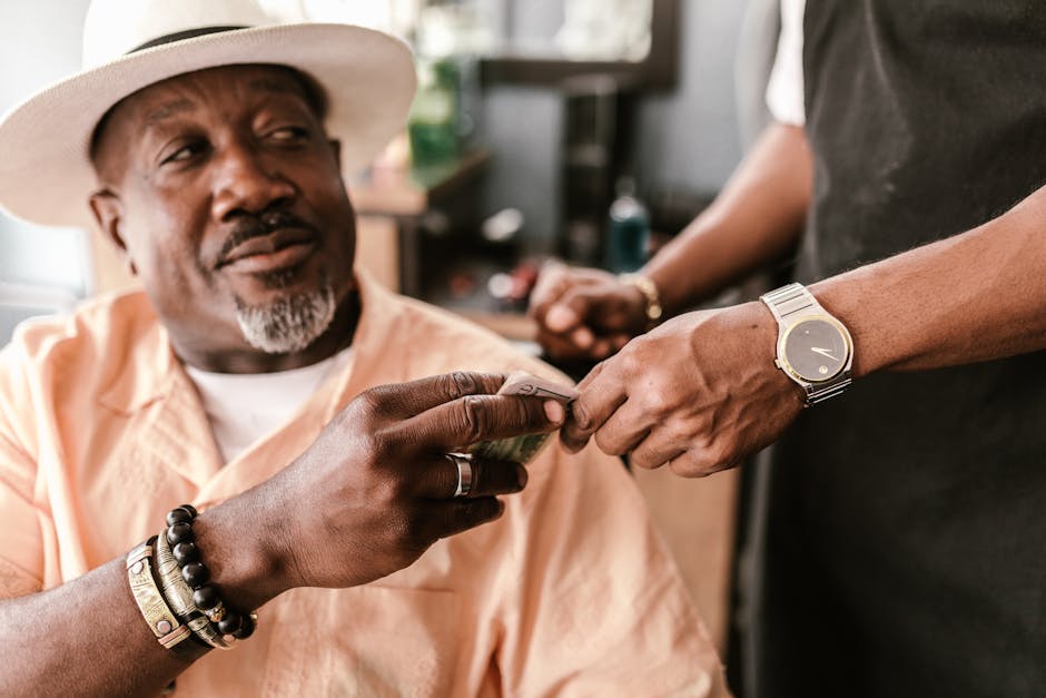 Senior man with a hat giving a tip to a young barber indoors, wearing bracelets and a watch.