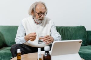 Senior man consulting doctor online, holding medicine and glass indoors.