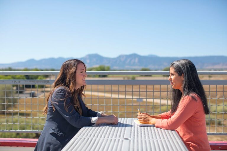 Two women discussing work at a table outdoors with a scenic mountain view.