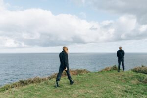 Two people in dark clothing stand on a grassy cliff edge overlooking the ocean under a cloudy sky.