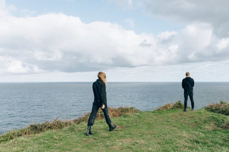 Two people in dark clothing stand on a grassy cliff edge overlooking the ocean under a cloudy sky.