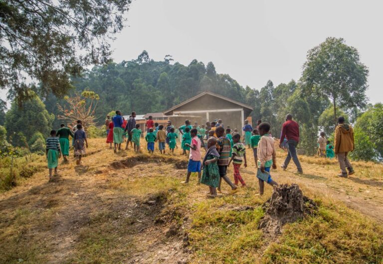 Group of children walking towards a school in a rural landscape surrounded by lush greenery.