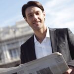 Smiling businessman in suit reads newspaper outside on a sunny day.