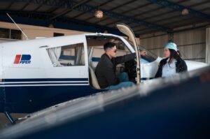 Two individuals conversing by a small aircraft in a hangar, suggesting aviation and teamwork.