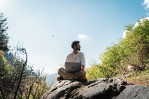 A man sits on a rock outdoors working on a laptop surrounded by lush greenery.