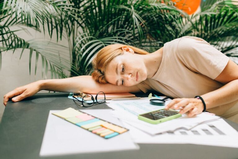 A tired woman calculating her taxes at a desk covered with papers and a smartphone.