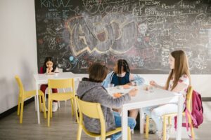 Children sitting in a school classroom having lunch with a chalkboard full of doodles behind them.